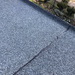 Jakub Markuszewski - Szczegółowe ujęcie pęknięcia na szorstkiej powierzchni dachu pokrytego drobnym kruszywem, z widocznym ogrodem i ogrodzeniem w tle.