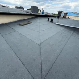 Wykończony dach pokryty papą termozgrzewalną w ciemnoszarym kolorze, widoczne kominy i lukarna. Dach po remoncie.