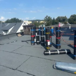 Bitumiczne rolki na płaskim dachu w trakcie prac dekarskich, widoczne narzędzia i preparaty. W tle zabudowa miejska i zieleń.