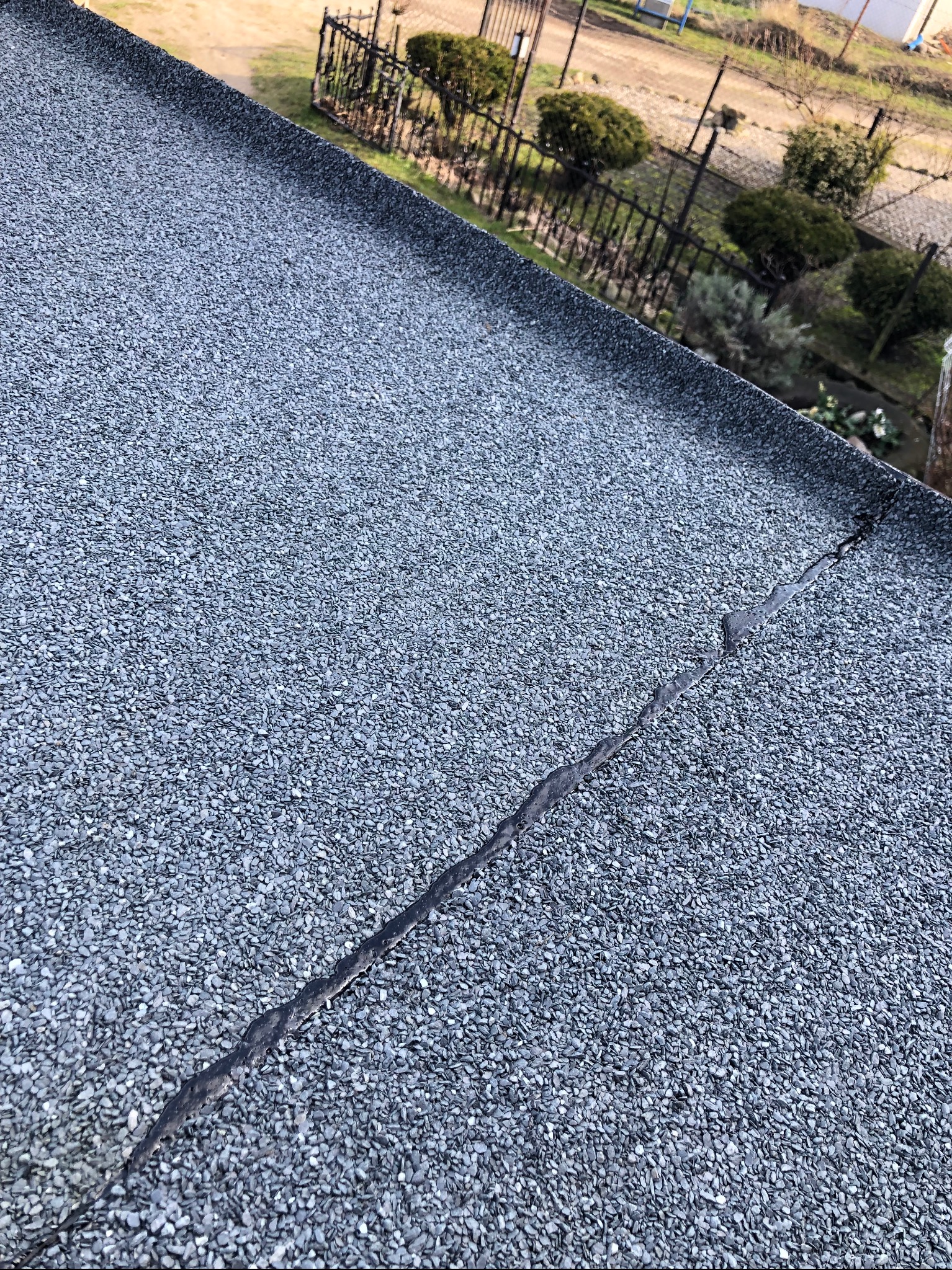 Szczegółowe ujęcie pęknięcia na szorstkiej powierzchni dachu pokrytego drobnym kruszywem, z widocznym ogrodem i ogrodzeniem w tle.