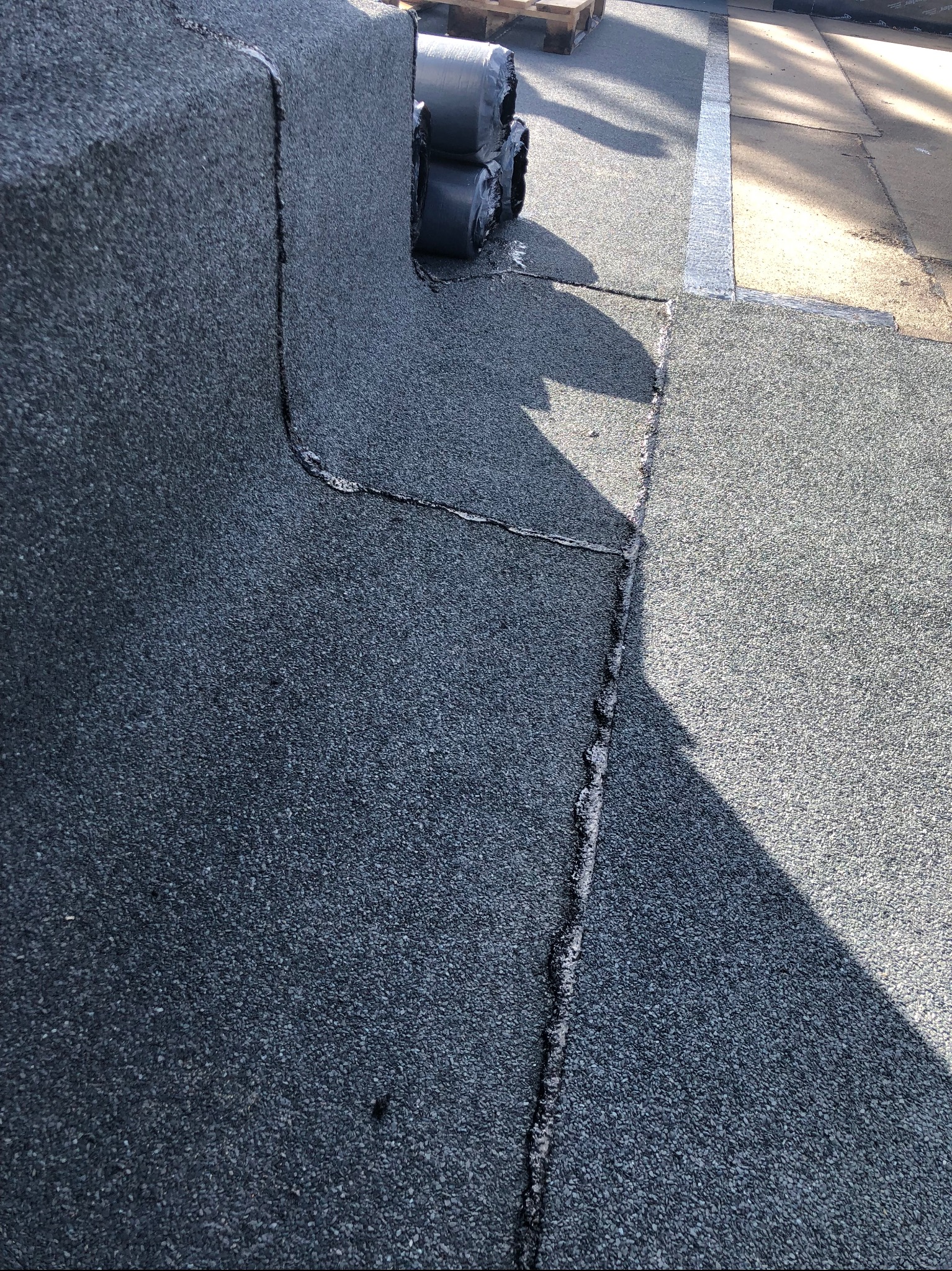 Fragment dachu pokrytego papą z widocznymi łączeniami i rolkami materiału na palecie w tle, oświetlony naturalnym światłem.