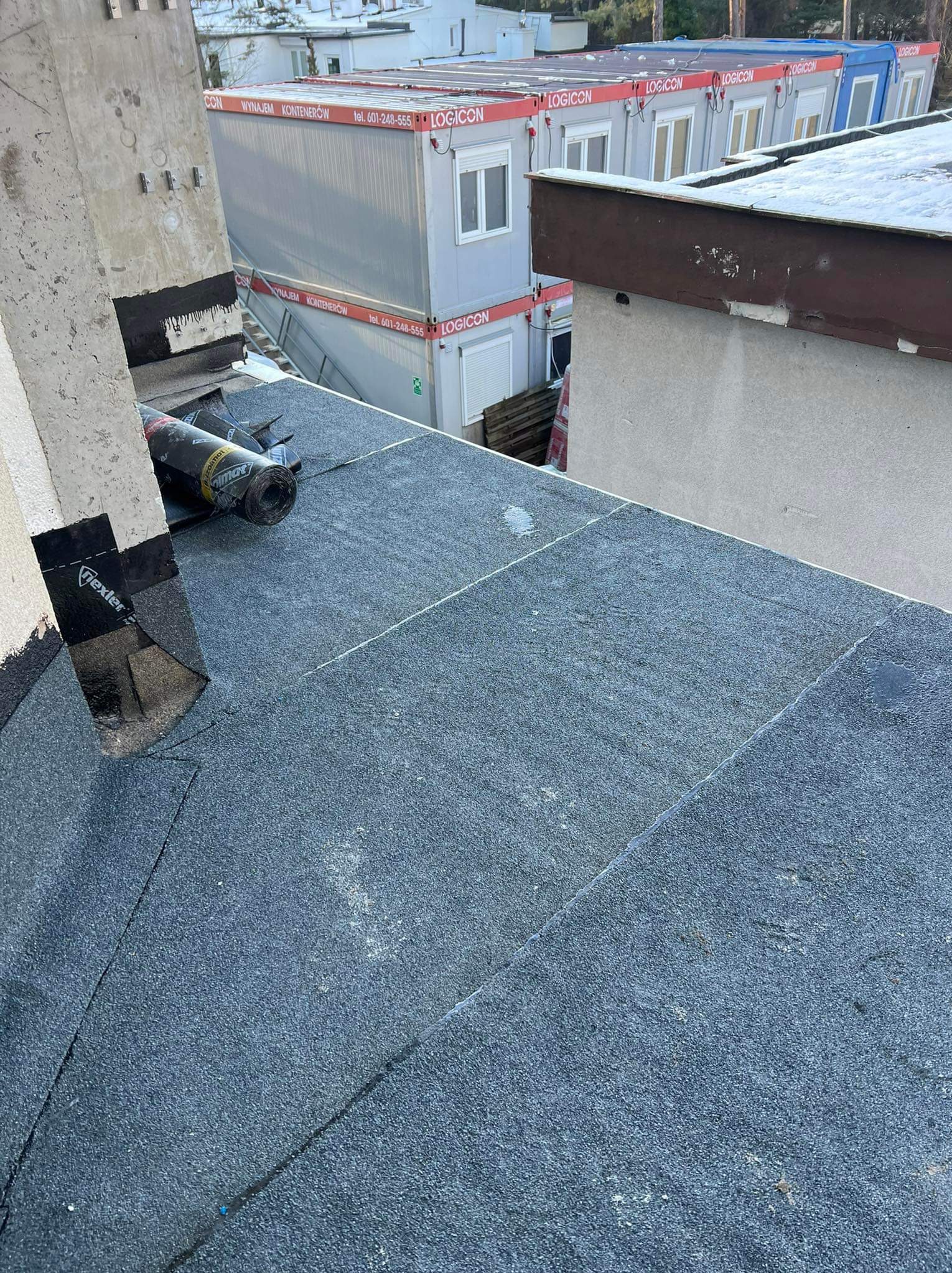 Fragment dachu pokrytego papą z widocznym fragmentem komina i rolką papy. W tle kontenery biurowe.