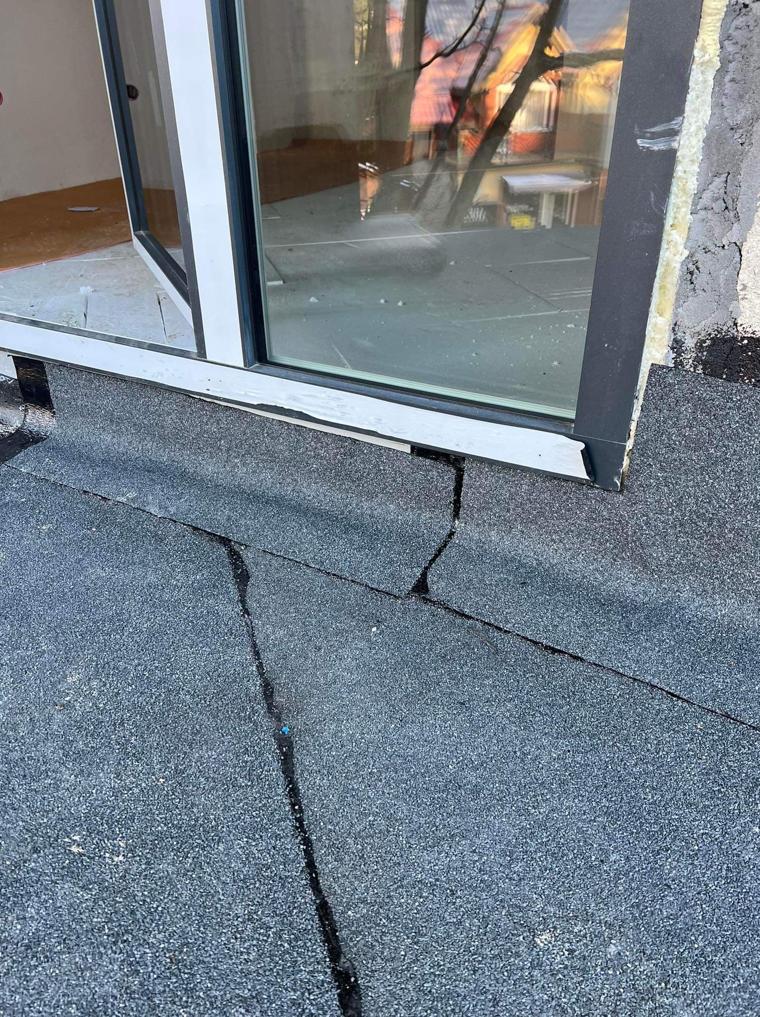 Pęknięcia na papie dachowej przy oknie dachowym. Widoczne uszkodzenia i nieszczelności warstwy hydroizolacyjnej dachu płaskiego.