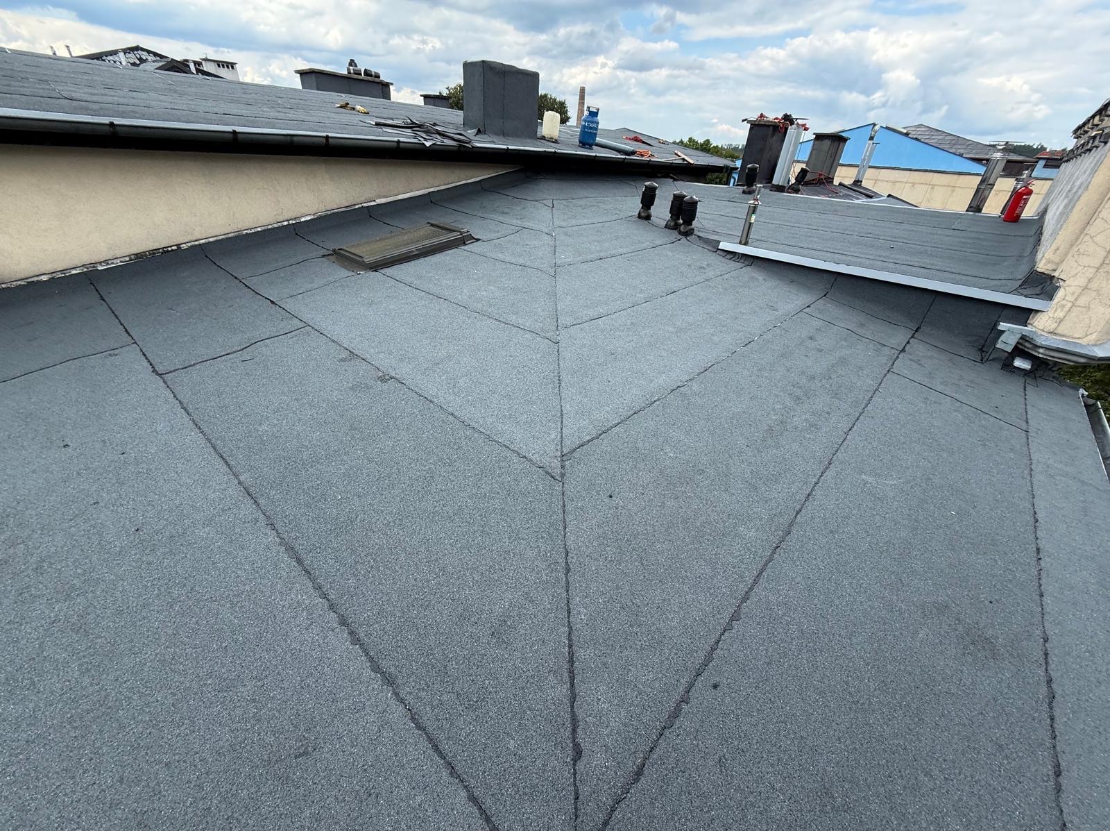 Wykończony dach pokryty papą termozgrzewalną w ciemnoszarym kolorze, widoczne kominy i lukarna. Dach po remoncie.