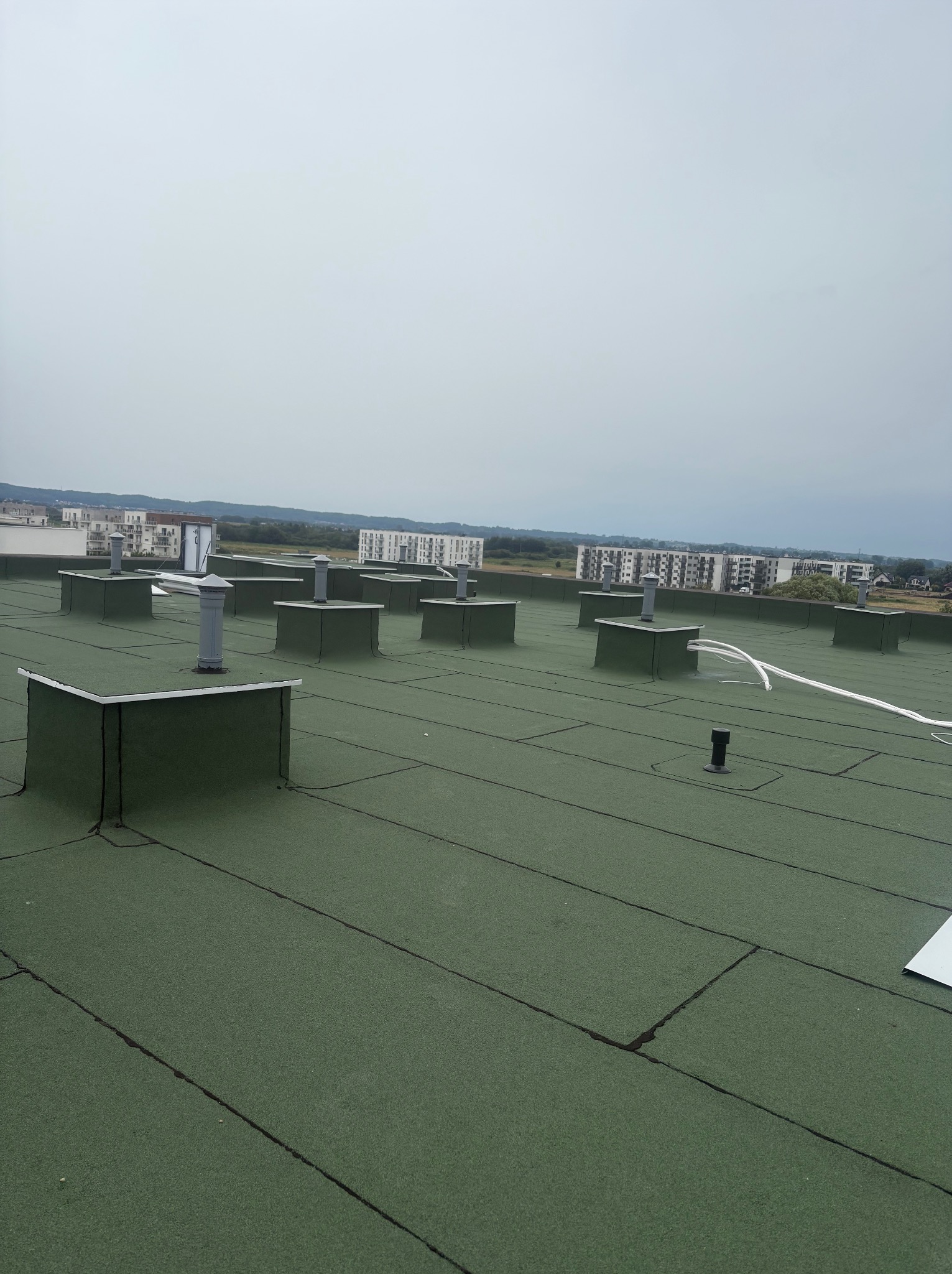 Zielony dach z licznymi kominkami wentylacyjnymi na tle zabudowy miejskiej pod pochmurnym niebem. Widoczne łączenia papy bitumicznej.