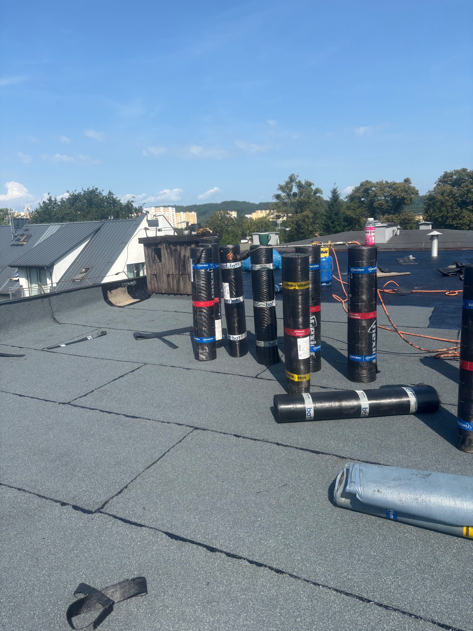 Bitumiczne rolki na płaskim dachu w trakcie prac dekarskich, widoczne narzędzia i preparaty. W tle zabudowa miejska i zieleń.