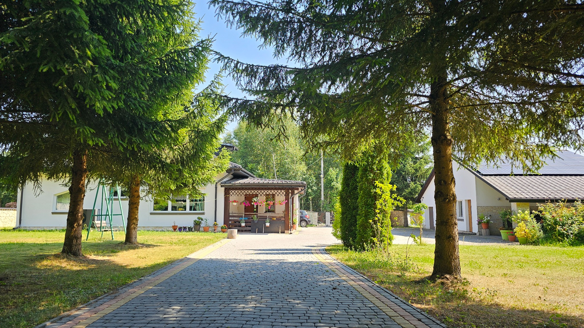 Szeroki, wybrukowany podjazd prowadzi do dwóch białych domów z brązowymi dachami, otoczonych zielonymi drzewami i krzewami, w słoneczny dzień.