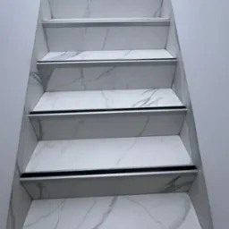 Schody wykończone białymi płytkami imitującymi marmur, z aluminiowymi listwami zabezpieczającymi krawędzie stopni.