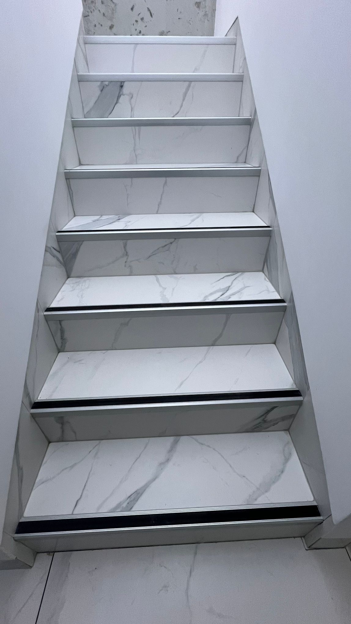 Schody wykończone białymi płytkami imitującymi marmur, z aluminiowymi listwami zabezpieczającymi krawędzie stopni.