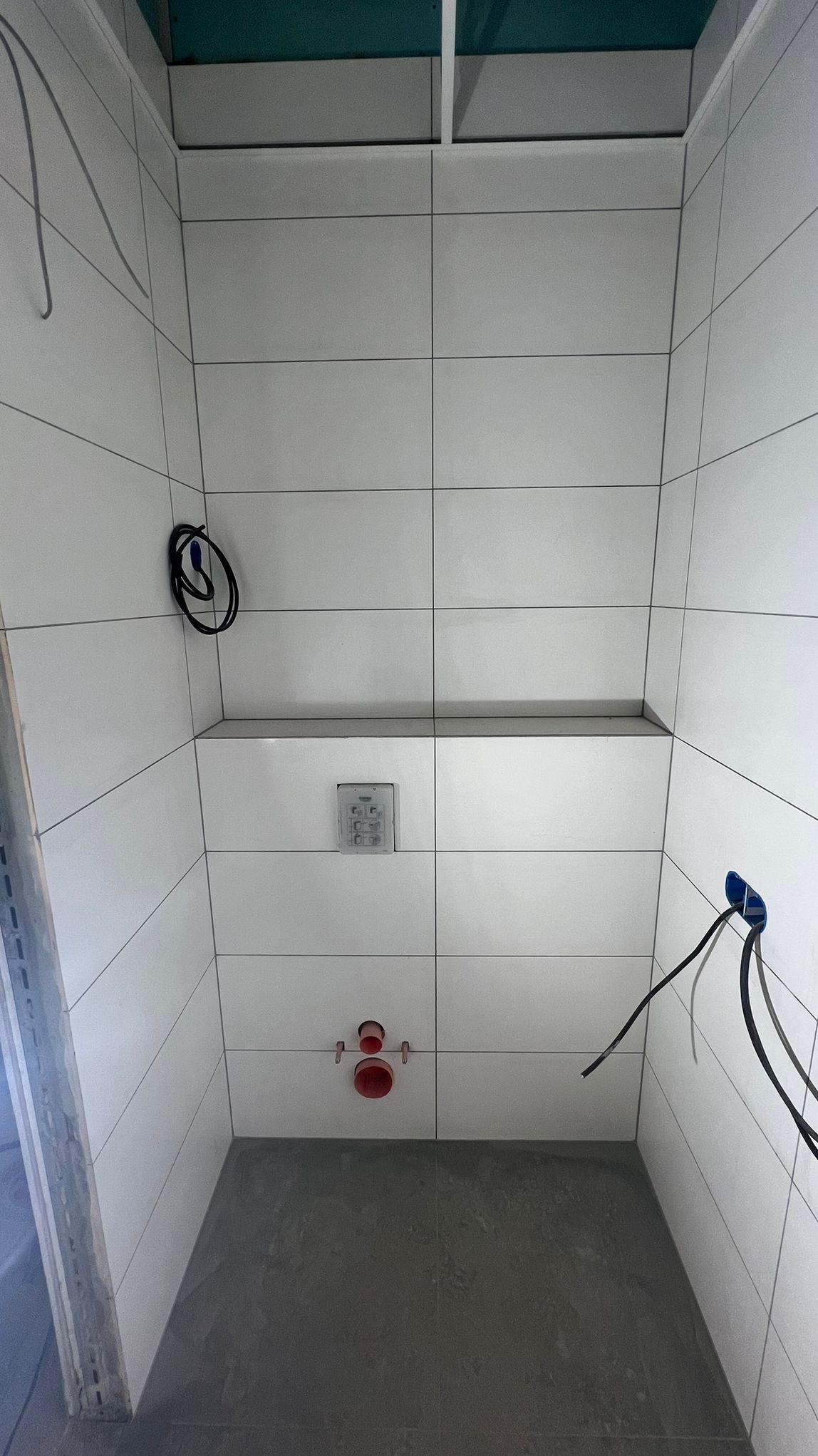Wnętrze łazienki w trakcie remontu: białe płytki ścienne z szarą fugą, szara podłoga, wystające rury instalacji wodnej i elektrycznej, widoczna instalacja podtynkowego systemu spłukiwania.