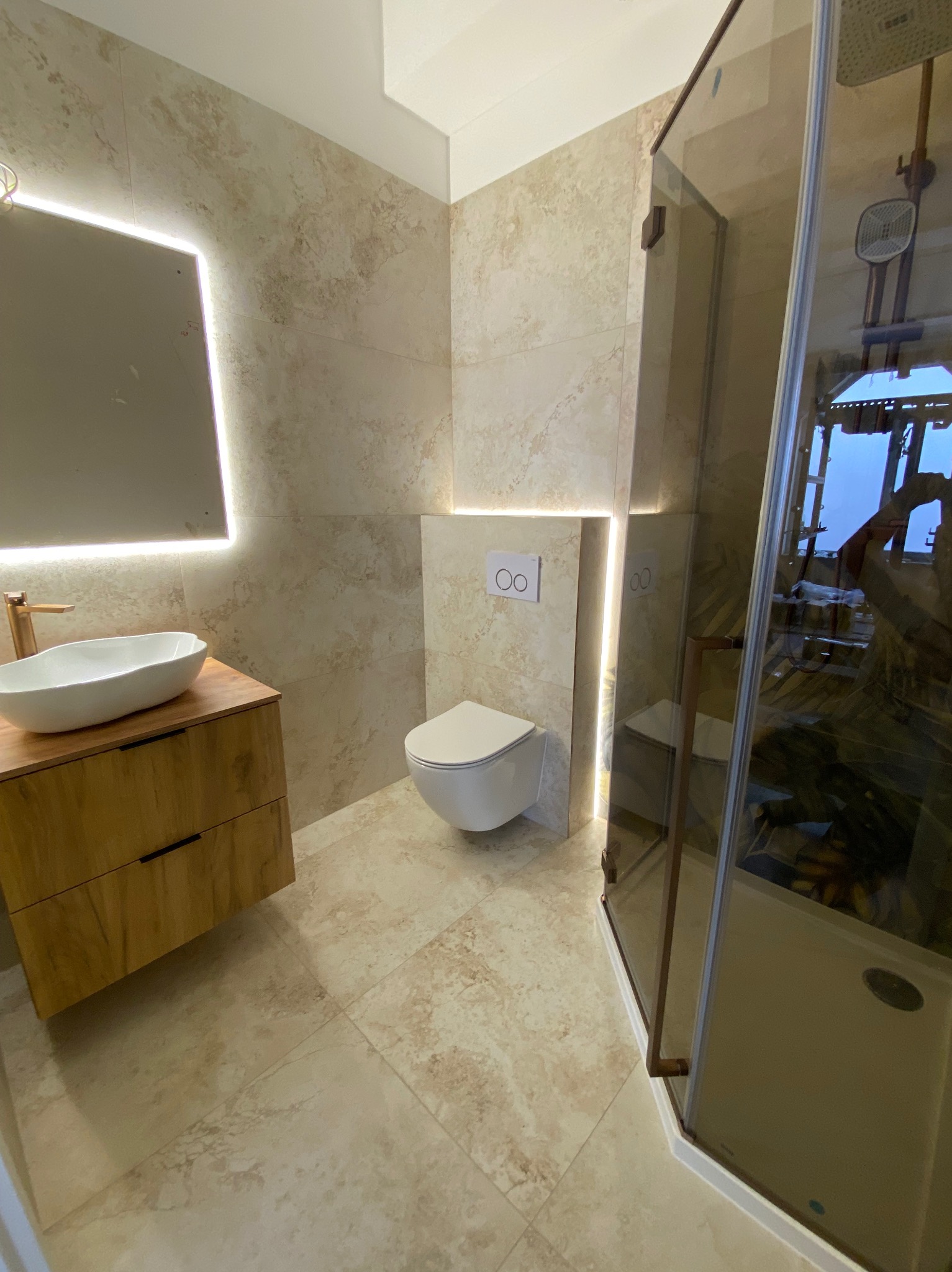 Nowoczesna łazienka w beżowych płytkach z podświetlanym lustrem, drewnianą szafką pod umywalką i kabiną prysznicową z brązowymi detalami.