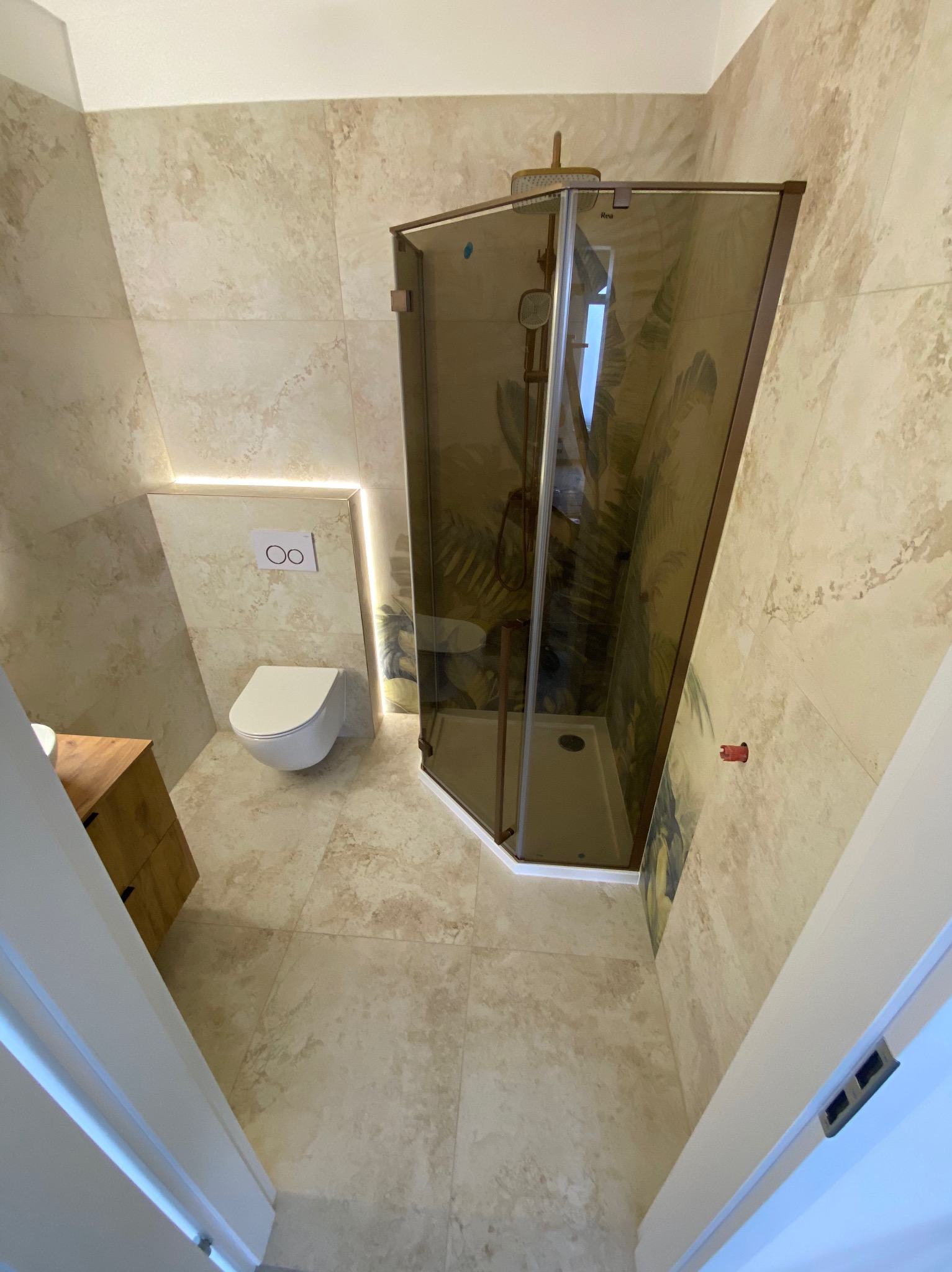 Wykończona łazienka z beżowymi płytkami imitującymi kamień, kabiną prysznicową z brązowymi ramami i toaletą podwieszaną z podświetleniem LED.