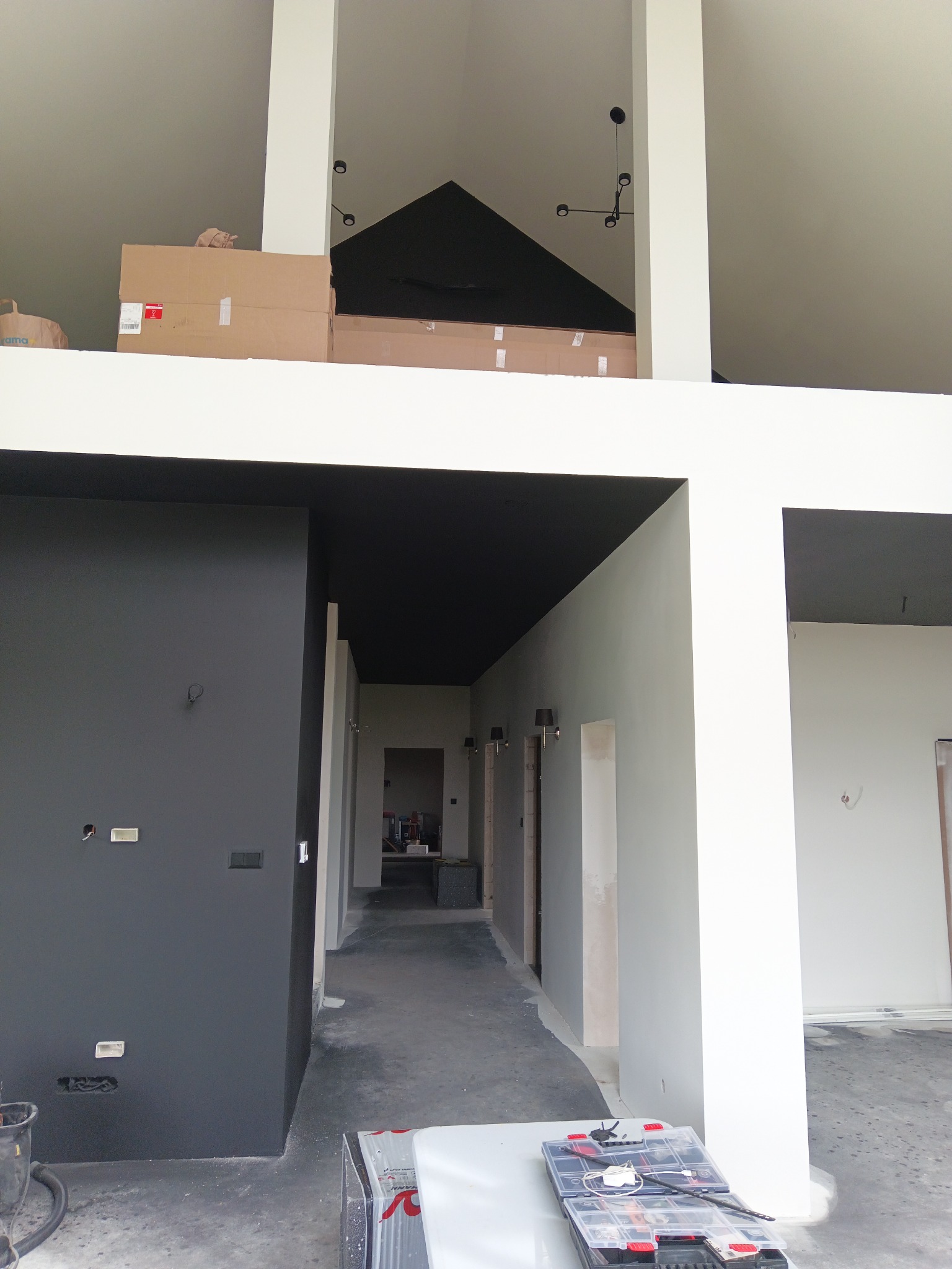 Surowe wnętrze dwupoziomowego domu w trakcie prac wykończeniowych: pomalowane na czarno sufity, szara ściana z widocznymi puszkami elektrycznymi, białe filary i ściany w odcieniu szarości, kartony...