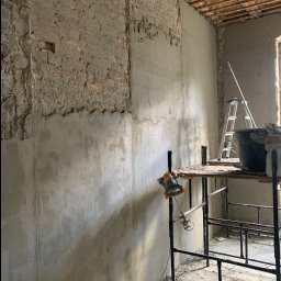 MATRE II - Ściana w trakcie prac remontowych: częściowo otynkowana, częściowo w stanie surowym z widoczną starą strukturą, rusztowanie z mieszadłem do zaprawy, drabina oparta o ścianę.