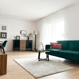 MATRE II - Jasne, przestronne wnętrze w Szczecinie z zieloną sofą, drewnianą podłogą i dywanem, oświetlone światłem dziennym wpadającym przez okno z białymi zasłonami.