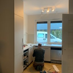 MATRE II - Montaż białych szafek kuchennych w nowym mieszkaniu, widok z tyłu na instalatora klęczącego na parkiecie w jodełkę, narzędzia leżą obok, za oknem zaparkowany biały bus