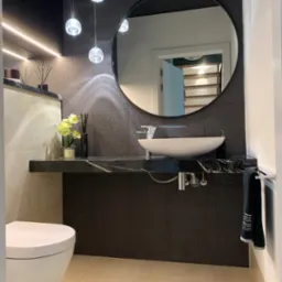 Nowoczesna łazienka z czarną marmurową umywalką, okrągłym lustrem, wiszącymi lampami kulami i beżowymi płytkami podłogowymi.