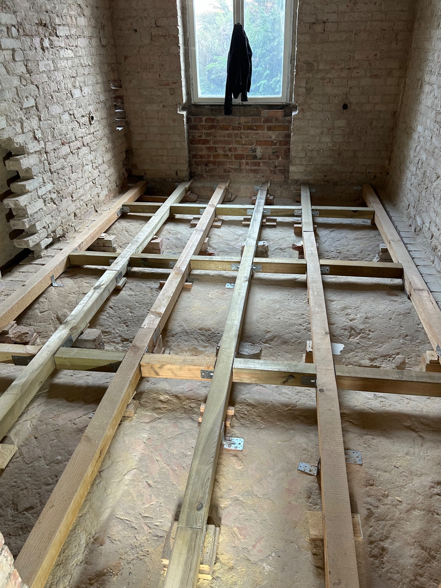 Konstrukcja drewniana pod podłogę na ceglanej posadzce w remontowanym pomieszczeniu z odsłoniętą cegłą i oknem, widoczne metalowe łączniki.