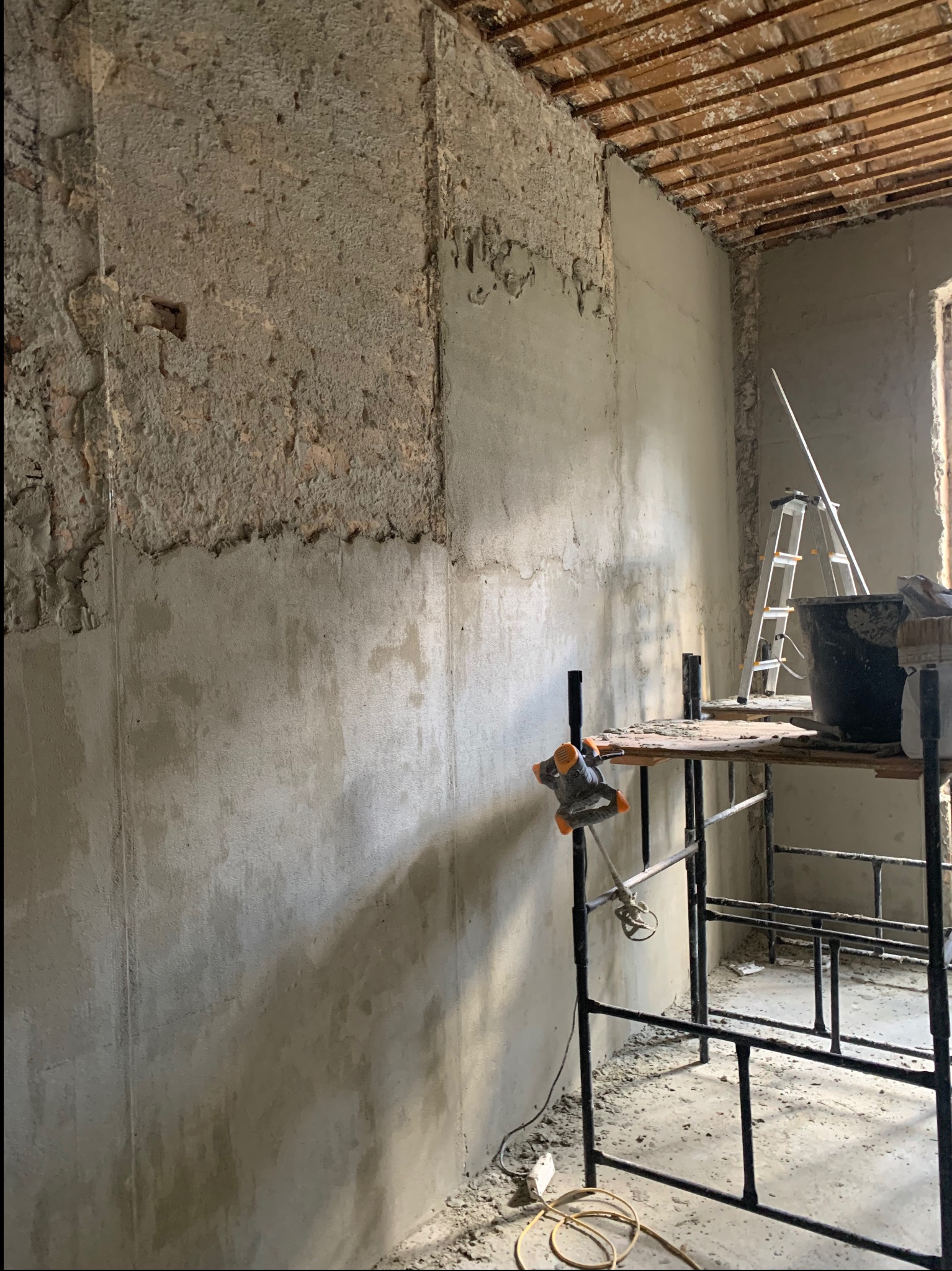 Ściana w trakcie prac remontowych: częściowo otynkowana, częściowo w stanie surowym z widoczną starą strukturą, rusztowanie z mieszadłem do zaprawy, drabina oparta o ścianę.