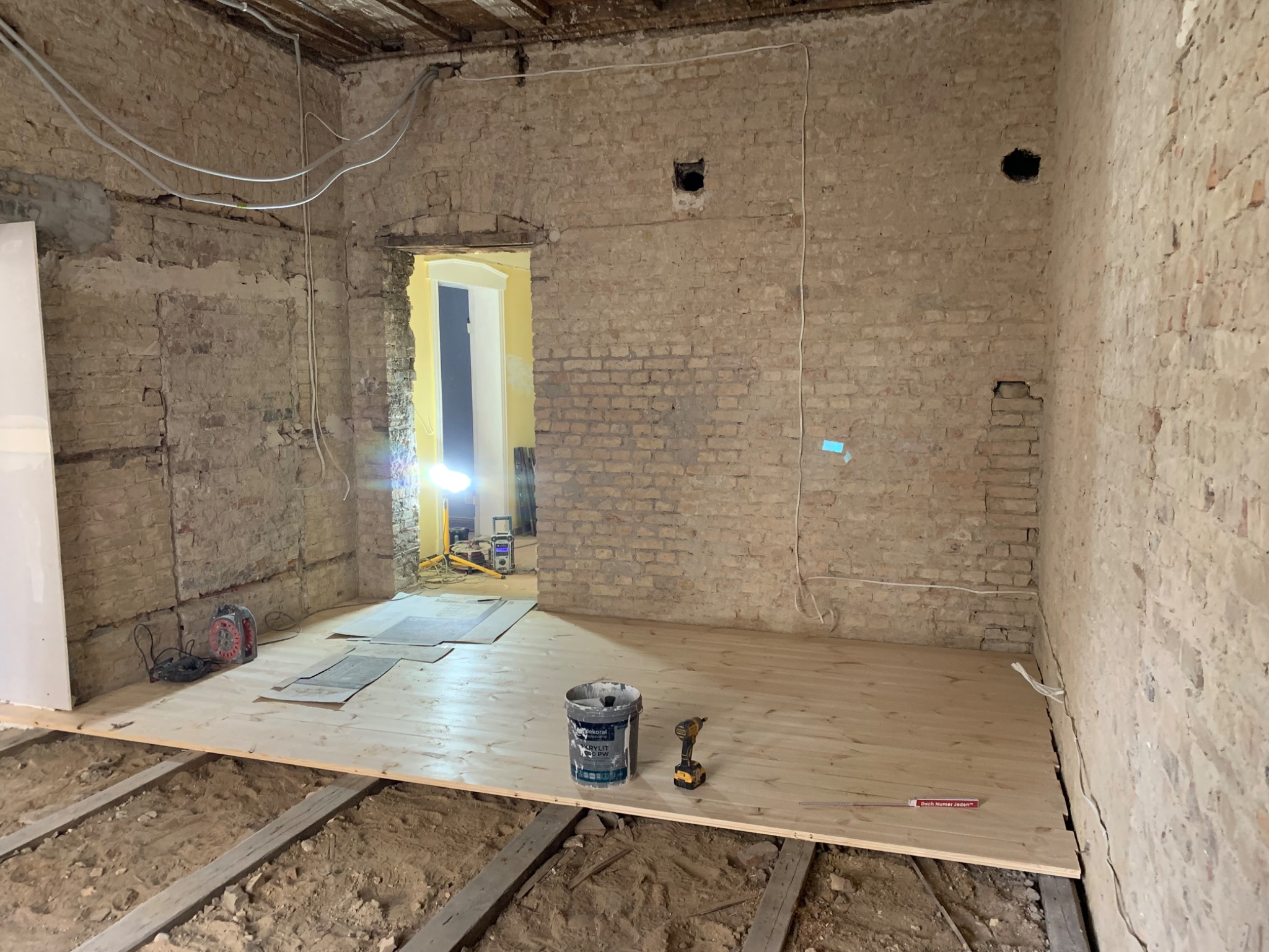 Pokój w trakcie remontu z surowymi ceglanymi ścianami, częściowo ułożoną drewnianą podłogą na legarach, widoczne narzędzia i puszka z farbą.