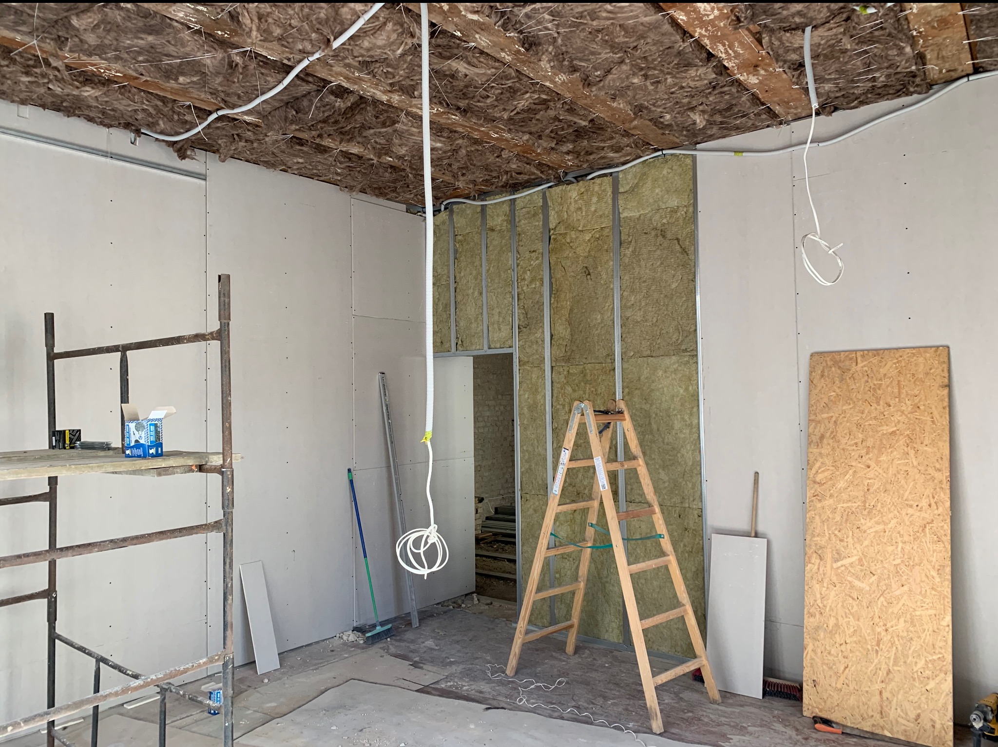 Wnętrze w trakcie prac remontowych: ściany częściowo obłożone płytami gipsowo-kartonowymi, izolacja z wełny mineralnej w metalowym stelażu, widoczne kable elektryczne, drabina i rusztowanie.