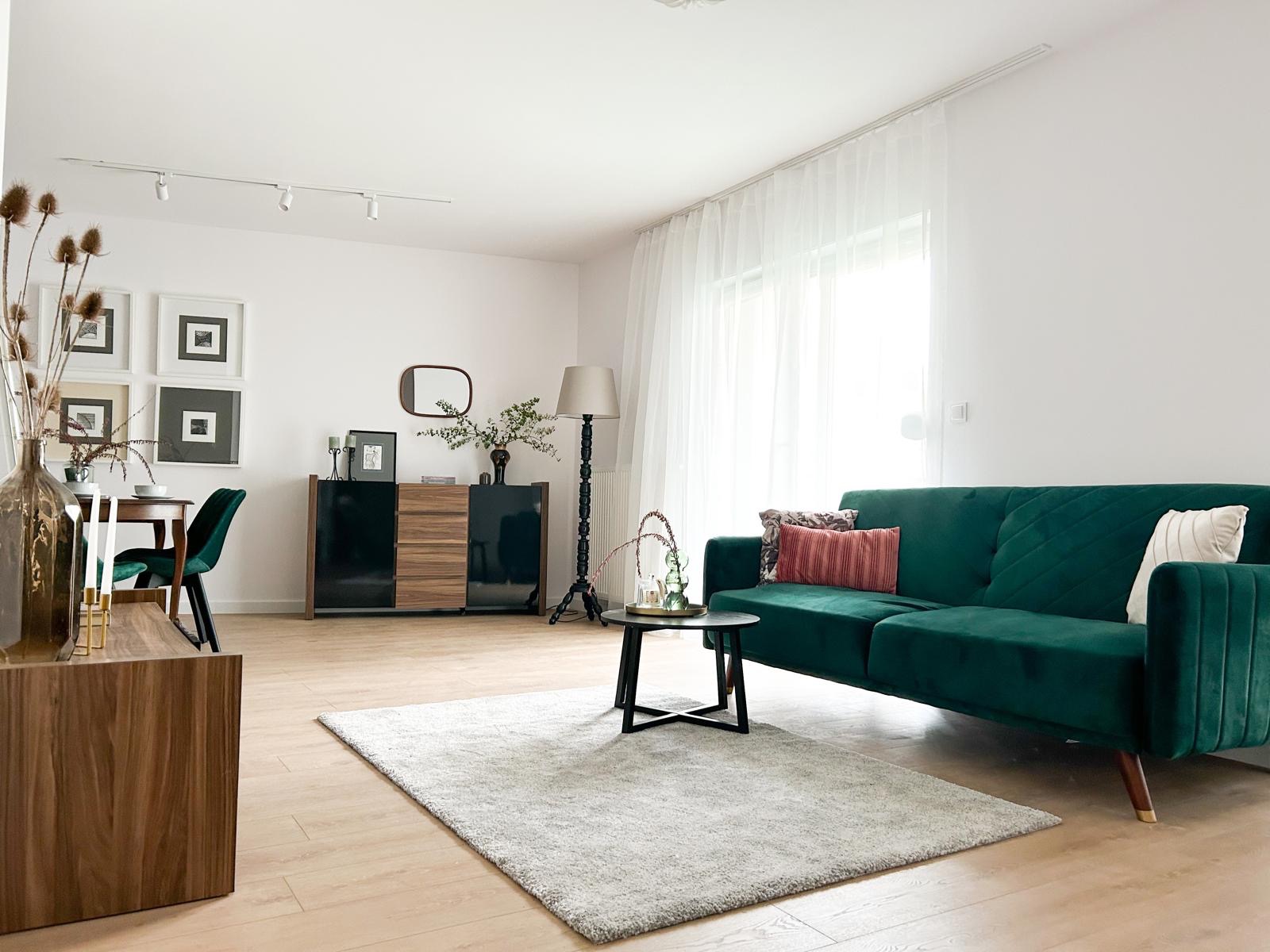 Jasne, przestronne wnętrze w Szczecinie z zieloną sofą, drewnianą podłogą i dywanem, oświetlone światłem dziennym wpadającym przez okno z białymi zasłonami.