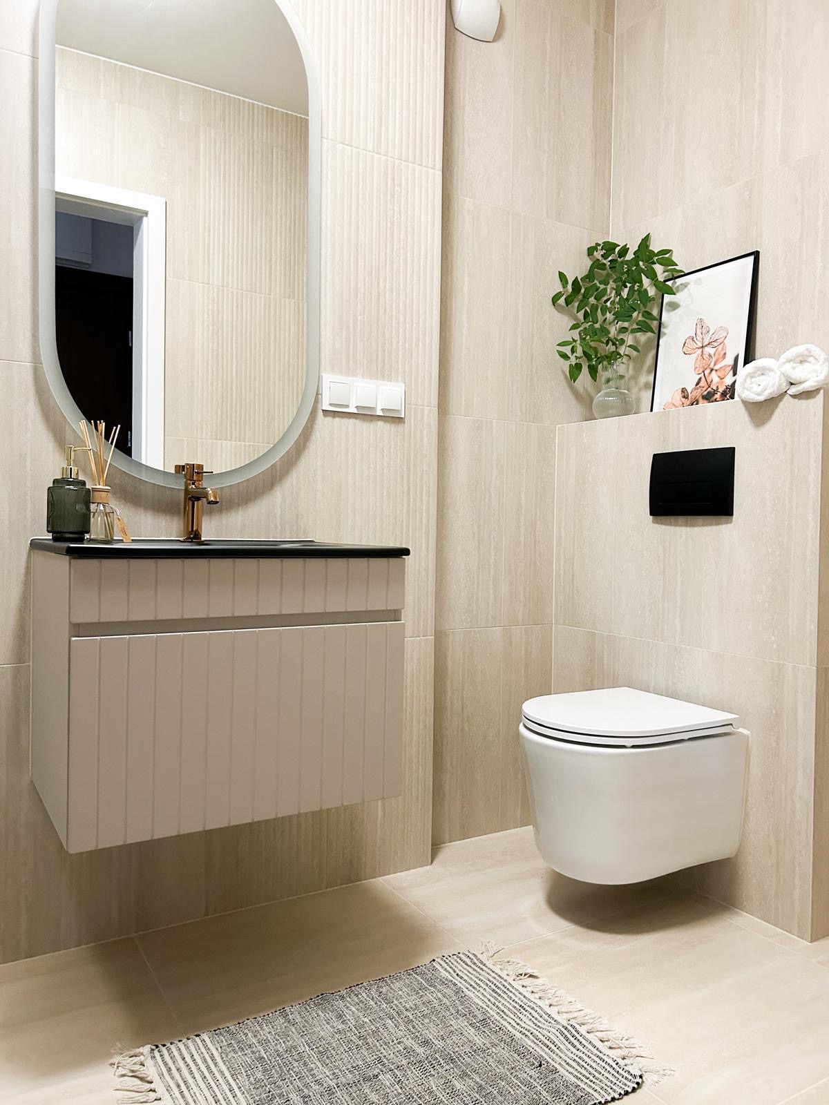Nowoczesna łazienka z beżowymi płytkami, podwieszaną toaletą, szafką pod umywalką z dekoracyjnymi frezami i owalnym lustrem, uzupełniona o dywanik w paski i roślinne dekoracje.