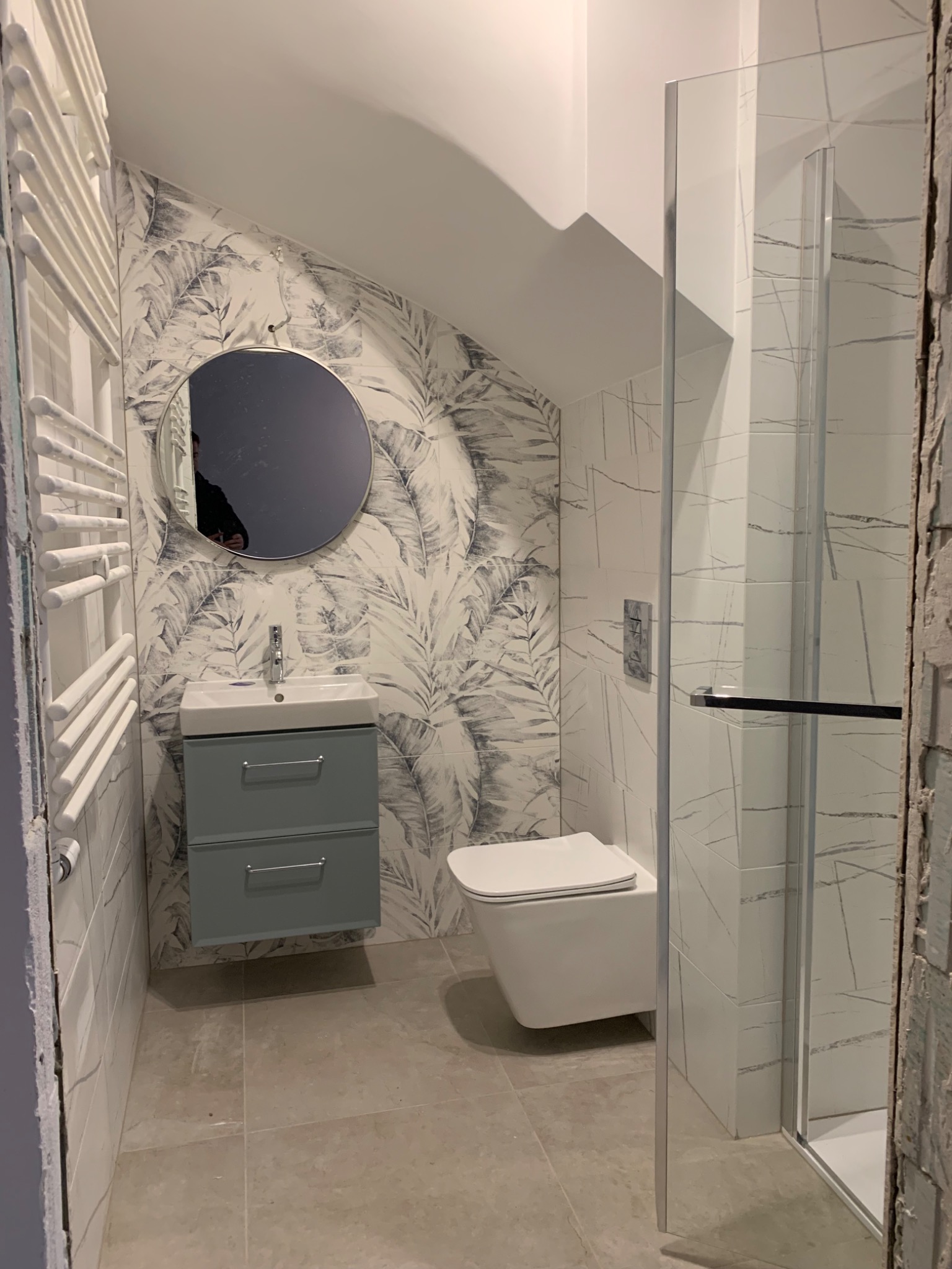 Nowoczesna łazienka na poddaszu z umywalką w kolorze gołębim, okrągłym lustrem, toaletą podwieszaną i kabiną prysznicową z czarnym uchwytem, wykończona płytkami z motywem liści i marmuru.