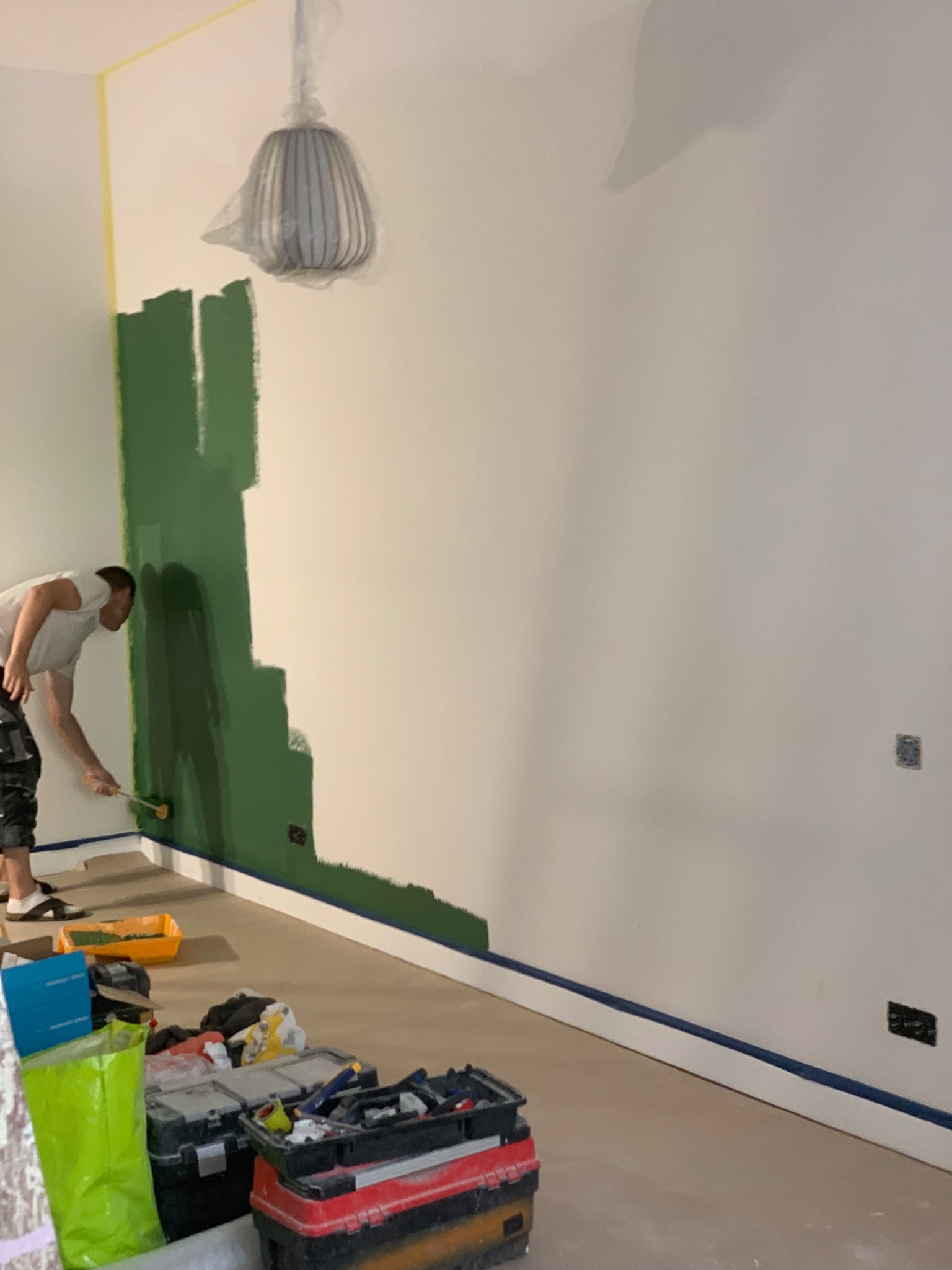 Mężczyzna maluje ścianę na zielono wałkiem, widoczne narzędzia malarskie i zabezpieczona podłoga taśmą malarską, lampa zabezpieczona folią.