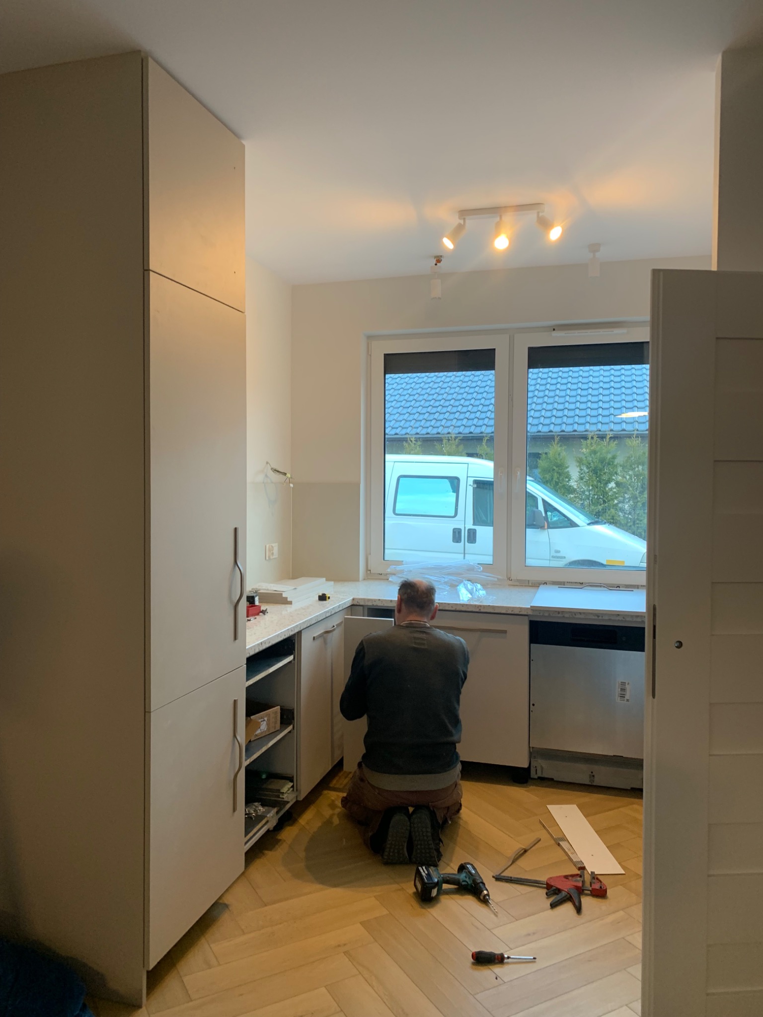 Montaż białych szafek kuchennych w nowym mieszkaniu, widok z tyłu na instalatora klęczącego na parkiecie w jodełkę, narzędzia leżą obok, za oknem zaparkowany biały bus
