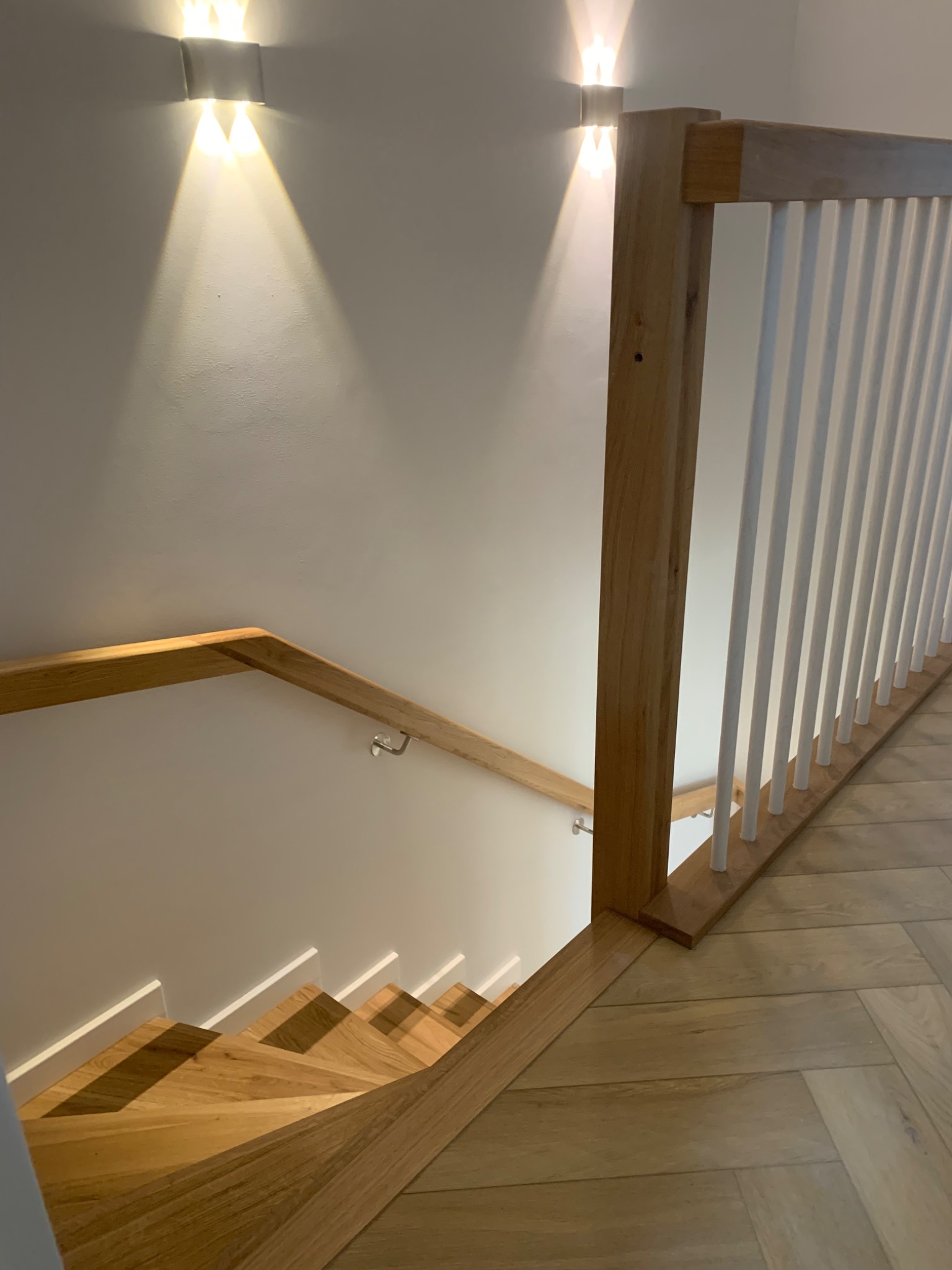 Drewniane schody z parkietem ułożonym w jodełkę, oświetlone ściennymi lampami z białymi poręczami i drewnianą balustradą.