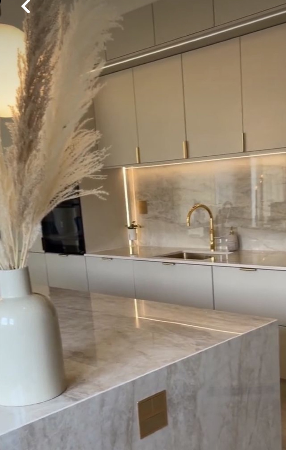 Nowoczesna kuchnia w odcieniach beżu i szarości z marmurowym blatem wyspowym i złotą armaturą, oświetlenie LED pod szafkami, w wazonie dekoracyjne trawy pampasowe.