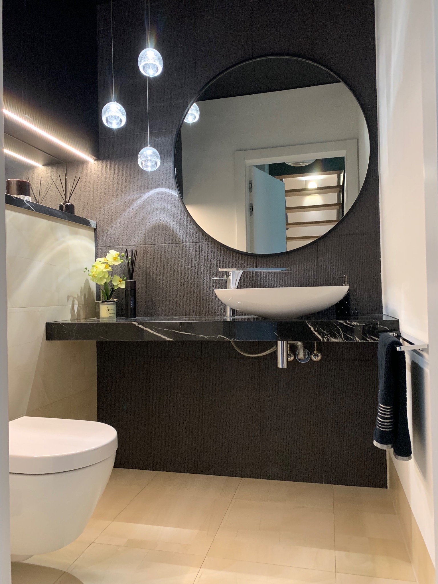 Nowoczesna łazienka z czarną marmurową umywalką, okrągłym lustrem, wiszącymi lampami kulami i beżowymi płytkami podłogowymi.