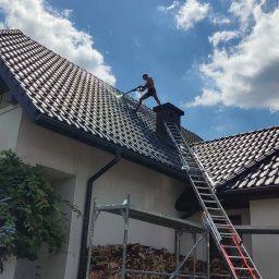 Adam Bysina - Mężczyzna bez koszulki myje dach z czarnej dachówki za pomocą myjki ciśnieniowej, widoczny komin, drabina oparta o dach, rusztowanie i stos drewna.