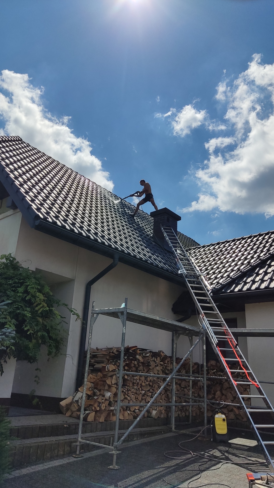 Mężczyzna bez koszulki myje dach z czarnej dachówki za pomocą myjki ciśnieniowej, widoczny komin, drabina oparta o dach, rusztowanie i stos drewna.