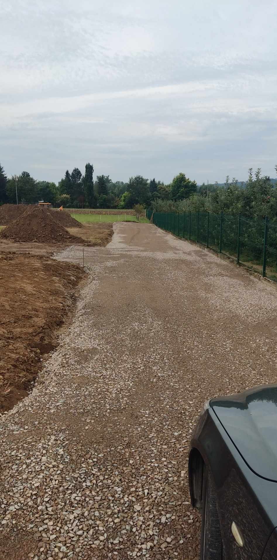 Widok na utwardzoną żwirową drogę dojazdową w trakcie budowy, z widocznymi hałdami ziemi, zielonym ogrodzeniem z siatki i fragmentem samochodu w prawym dolnym rogu.