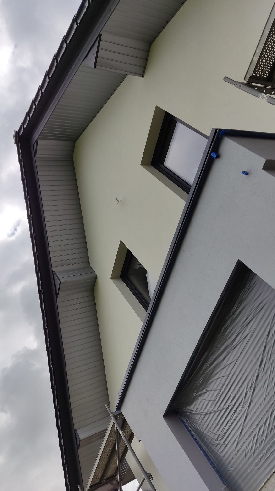 Ocieplona elewacja budynku z widocznymi oknami, fragmentem dachu i rusztowaniem, na tle pochmurnego nieba. Okno zabezpieczone folią malarską.