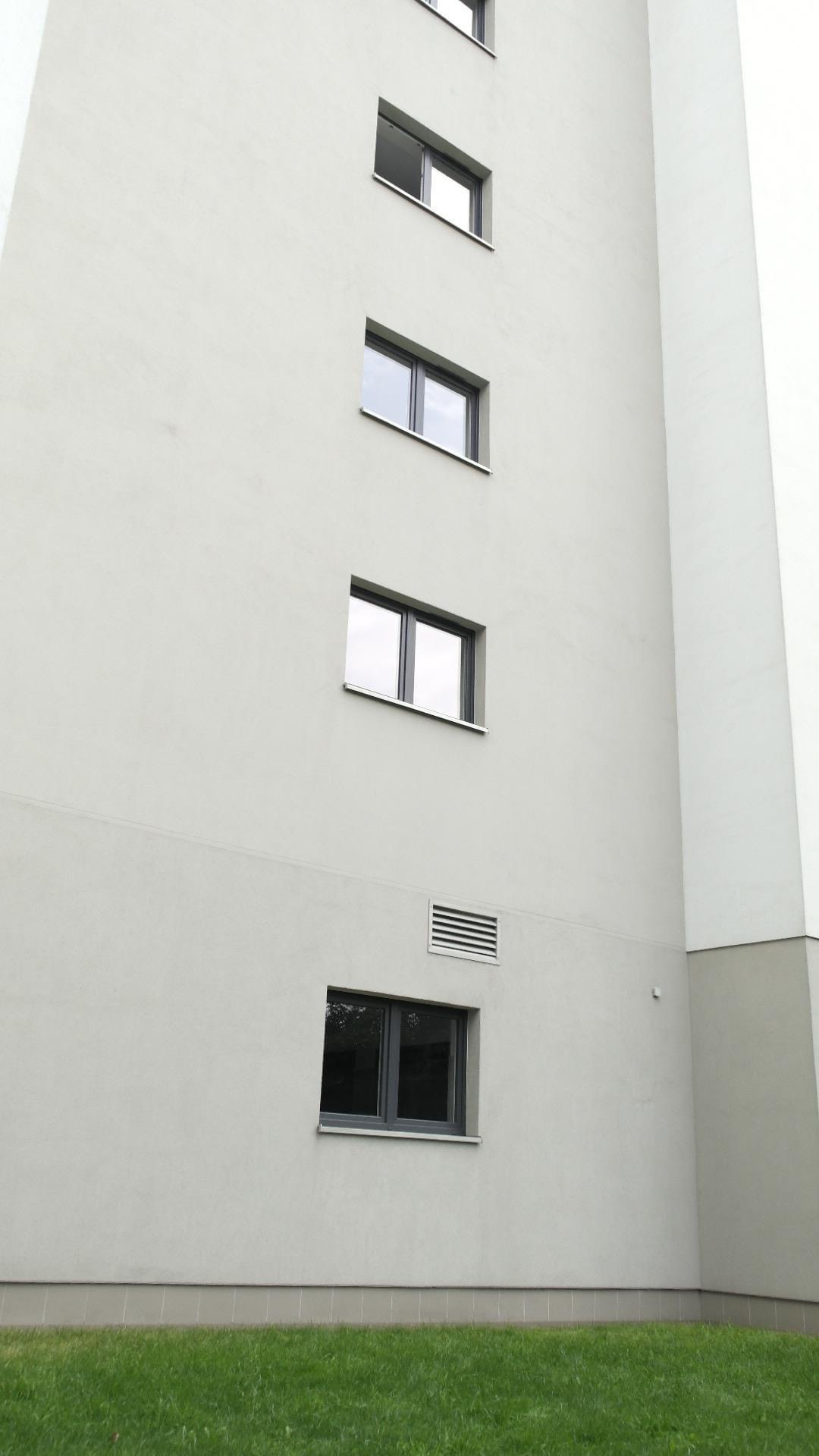Fragment elewacji budynku wielorodzinnego z oknami i kratką wentylacyjną, widok z dołu na tle trawnika.