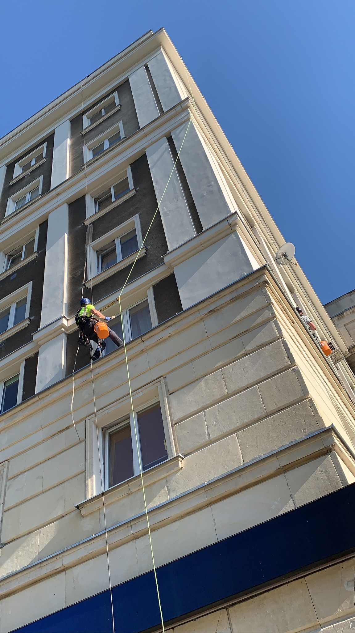 Pracownik alpinistyczny na linach myje okna wysokiego budynku z beżową elewacją i ciemnymi elementami, widok z dołu w słoneczny dzień.