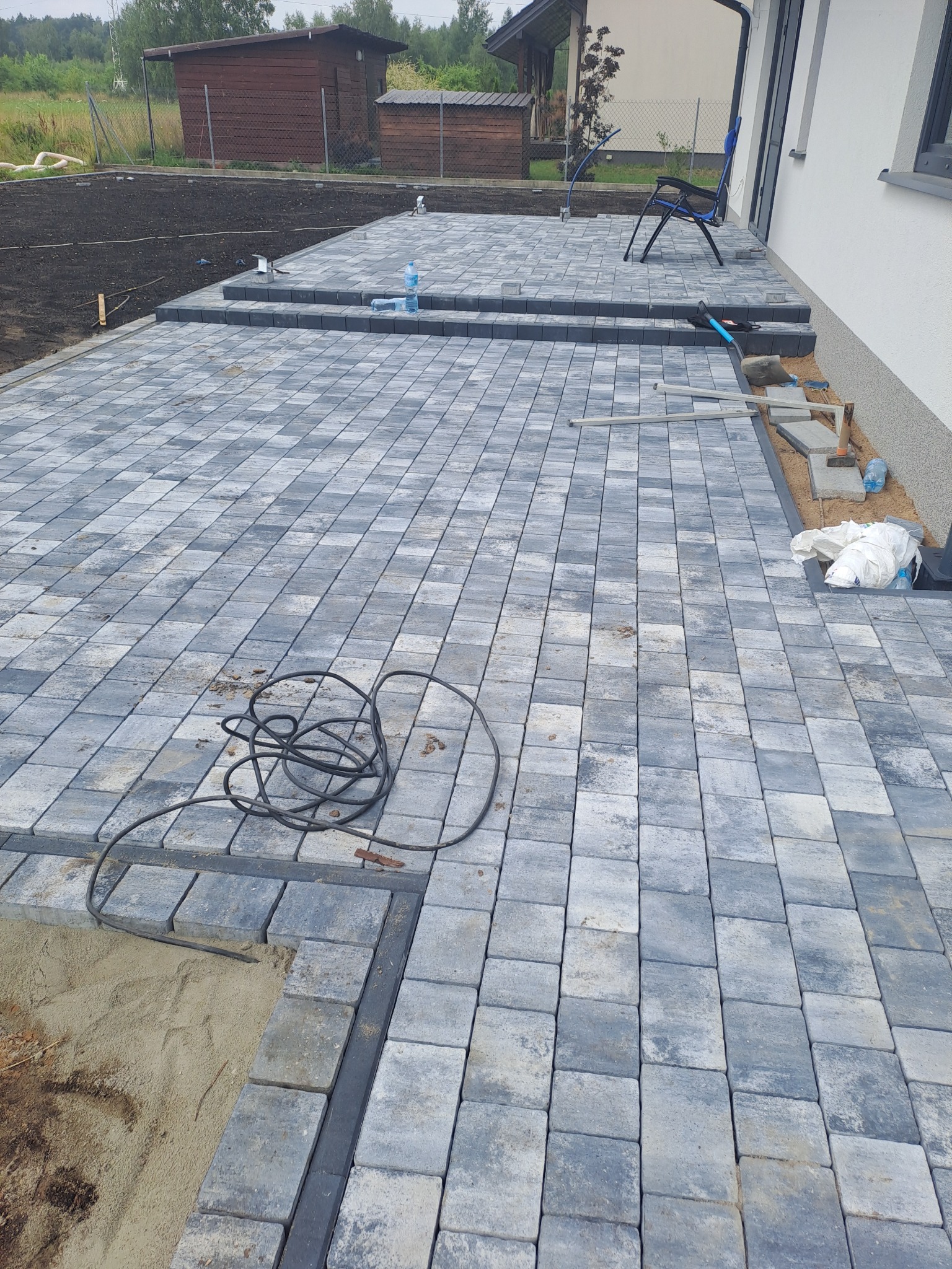 Świeżo ułożona, szara kostka brukowa w jodełkę na posesji, widoczny czarny kabel, butelka wody i narzędzia budowlane przy ścianie budynku.