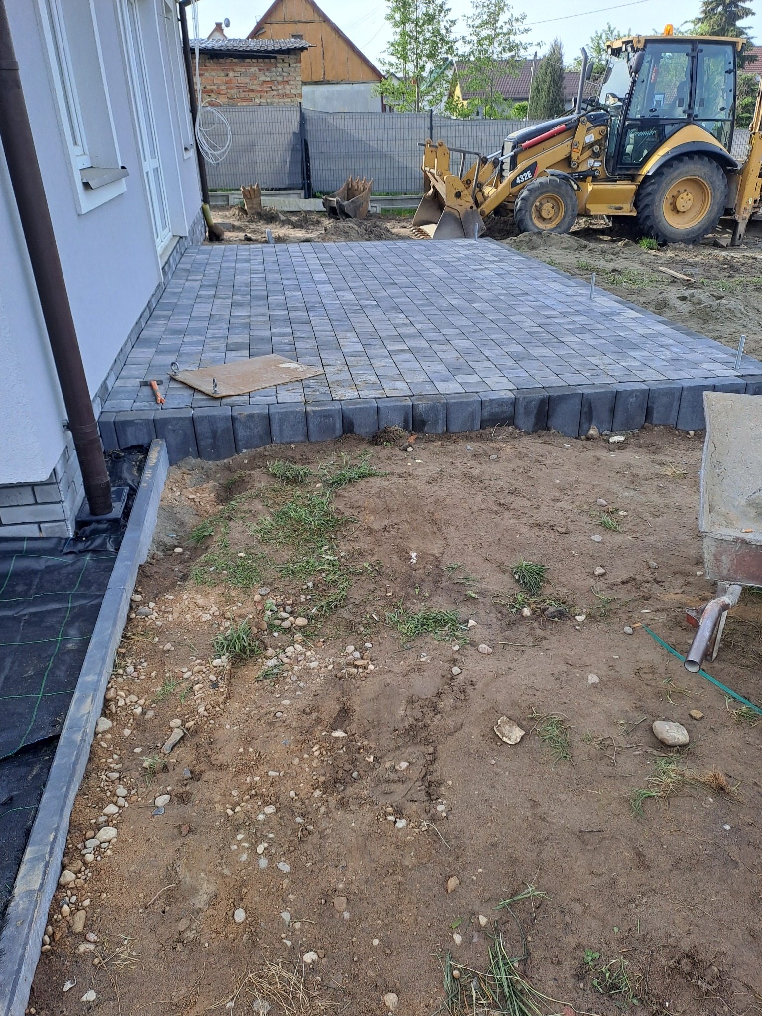 Częściowo ułożona szara kostka brukowa przy budynku, widoczny traktor z łyżką i teren przygotowany pod dalsze prace.