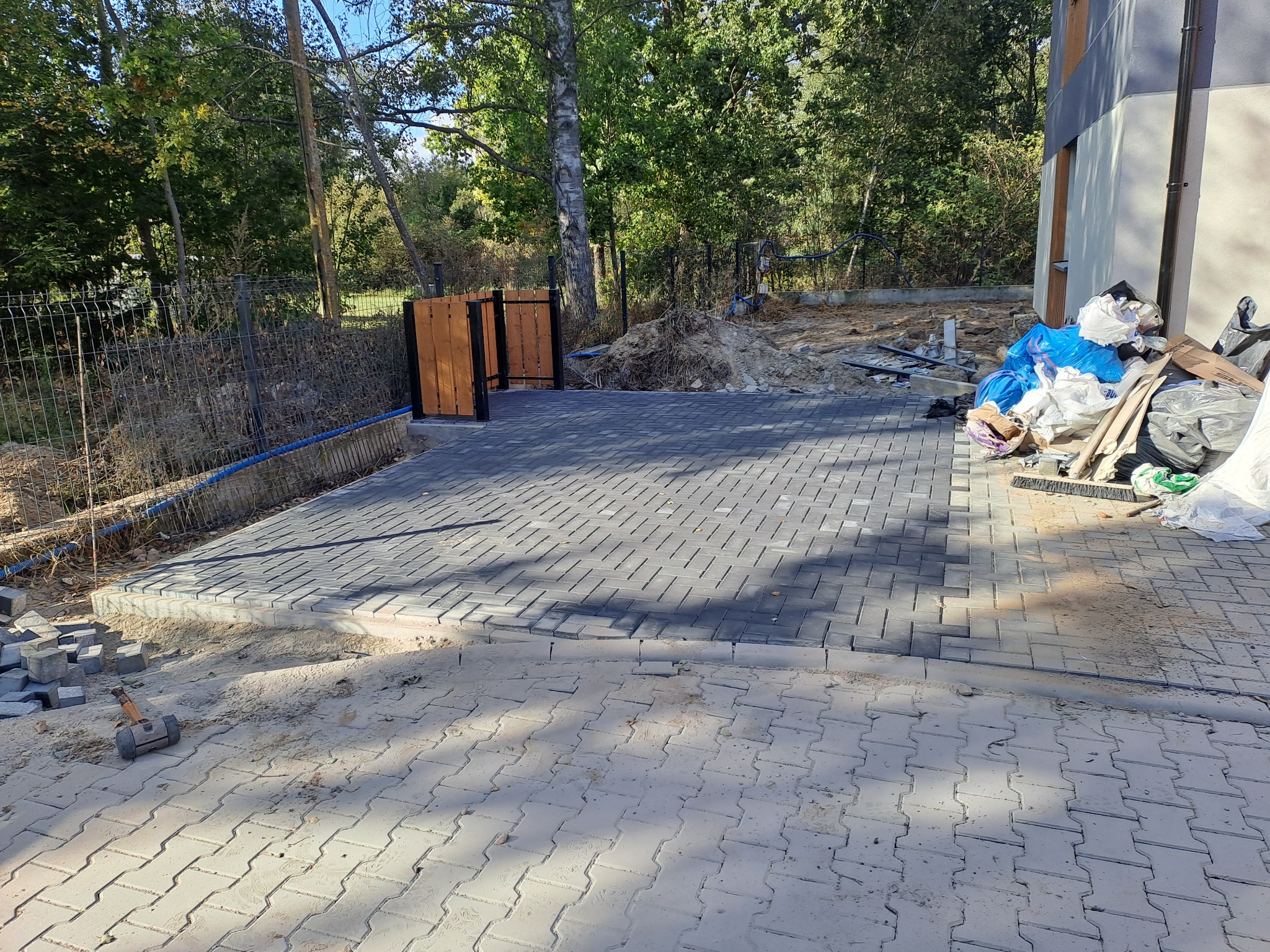 Układanie szarej kostki brukowej na posesji, widoczny gumowy młotek, obok domu i ogrodzenia, częściowo zagospodarowany teren.