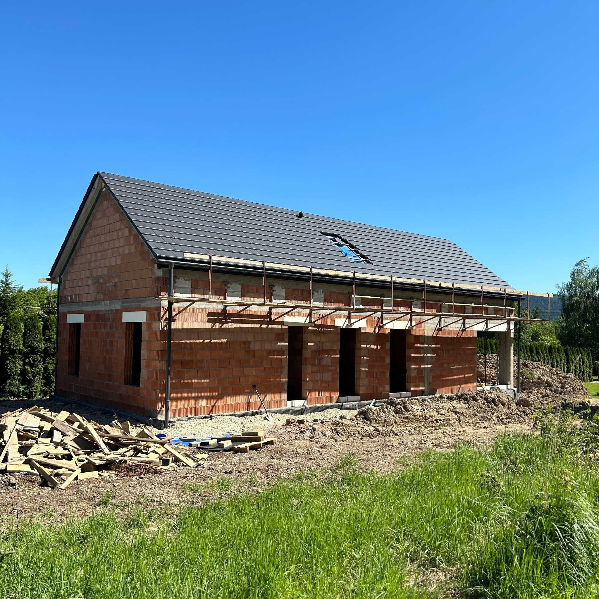 Budynek w stanie surowym otwartym z cegły, z częściowo położonym dachem z dachówki, otoczony zielenią i stertą drewna, widoczne rusztowania.