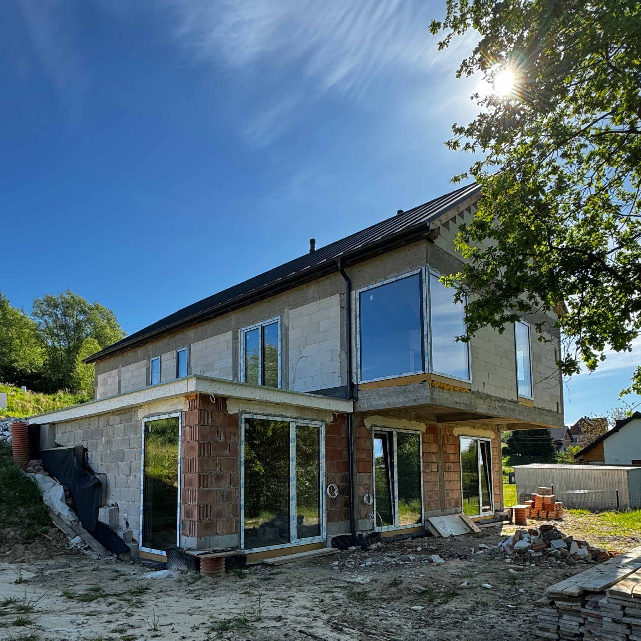 Niedokończony dwukondygnacyjny dom z dużymi oknami, widoczne pustaki i cegły, teren budowy z gruzem i materiałami, błękitne niebo z promieniami słońca.