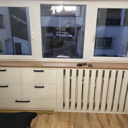 LookMeble - Zabudowa okna z białymi szafkami z czarnymi uchwytami i dekoracyjnym grzejnikiem. Blat drewniany, widok na podwórko z rowerem i samochodami.