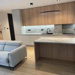 LookMeble - Nowoczesna kuchnia z drewnianymi frontami i wyspą, oświetlona lampą pierścieniową. Szary narożnik w tle. Minimalistyczny design z akcentami czerni.