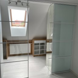 LookMeble - Szklana szafa wnękowa z przesuwnymi drzwiami w jasnym pokoju na poddaszu, z widocznym oknem dachowym i grzejnikiem zabudowanym w meblach.