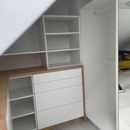 LookMeble - Biała szafa wnękowa z szufladami i półkami, dopasowana do skosu dachu, z drewnianym blatem. Widok z perspektywy wnętrza szafy, z fragmentem lustra po prawej.