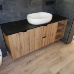 Drewniana szafka łazienkowa z czarnym blatem i owalną umywalką, widok z perspektywy podłogi, nowoczesny design, oświetlenie górne.