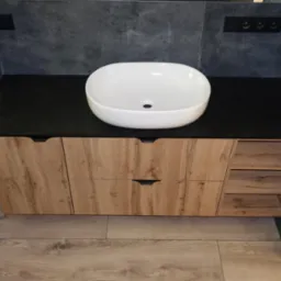 Nowoczesna szafka łazienkowa z blatem w kolorze czarnym i umywalką nablatową w kształcie owalnym. Fronty szafki w odcieniach drewna. Widoczne półki po prawej stronie.